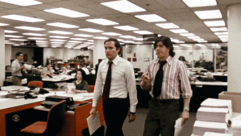 two men are walking in an office with a cbs logo on the cubicle