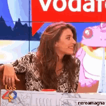 a woman sits at a table in front of a red sign that says vodafone