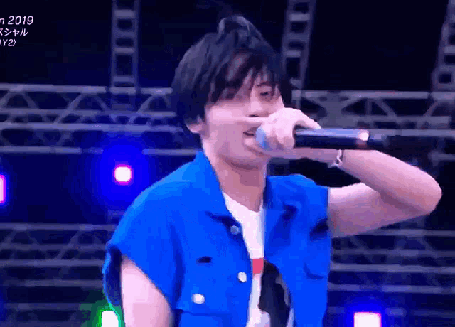 a man in a blue vest is singing into a microphone at a festival