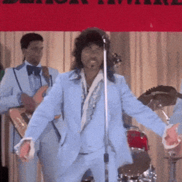 a man in a blue suit is singing into a microphone in front of a drum set .