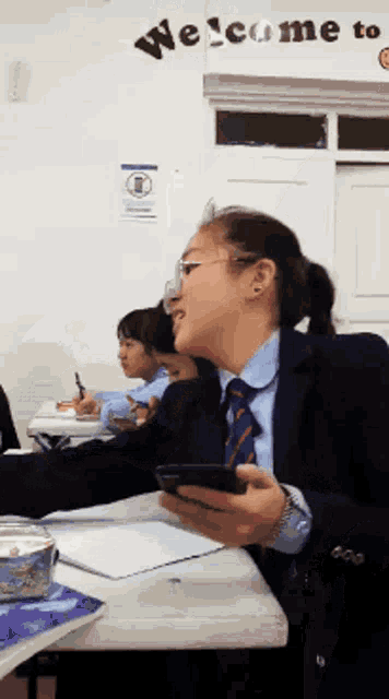 a woman in a suit and tie is sitting at a desk in front of a wall that says welcome to