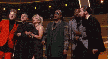 a group of people standing in front of microphones on a stage including a man in a green suit