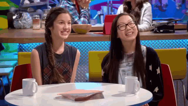 two girls are sitting at a table with cups and a book