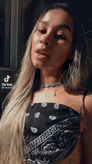 a woman wearing a black bandana top and a necklace has a tiktok sticker on her face