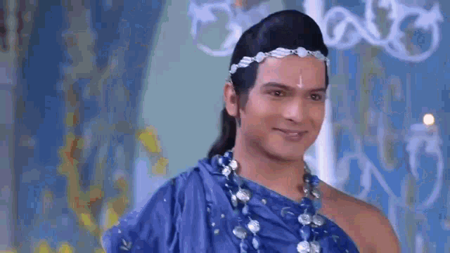 a man in a blue dress is smiling and wearing a headband and necklace .