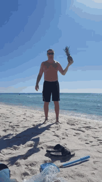 a shirtless man holds a pineapple in his hand on the beach