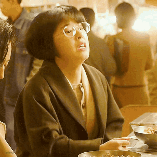 a woman wearing glasses sits at a table with a bowl of food in front of her