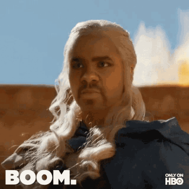a man with long blonde hair and a beard is standing in front of a fire and the word boom is above him .