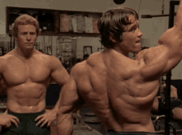 two shirtless men are standing next to each other in a gym and one is flexing his muscles