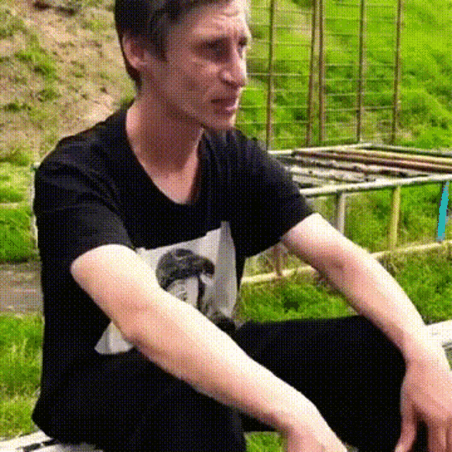 a man wearing a black shirt with a picture of a mushroom on it is sitting in the grass