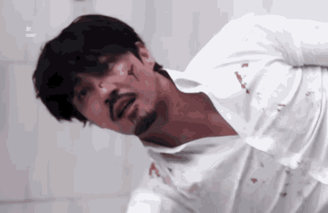 a man with a beard and blood on his face is wearing a white shirt and laying on the floor .