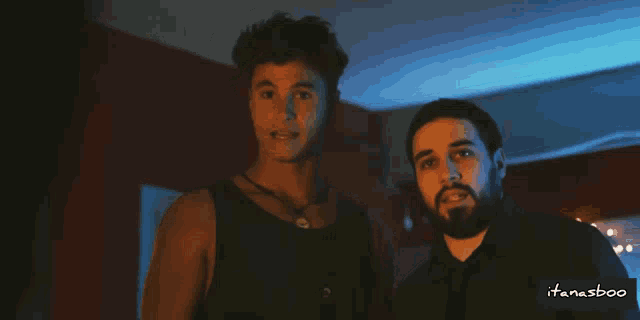 a man with a beard stands next to another man in a dark room with a watermark that says itanasboo