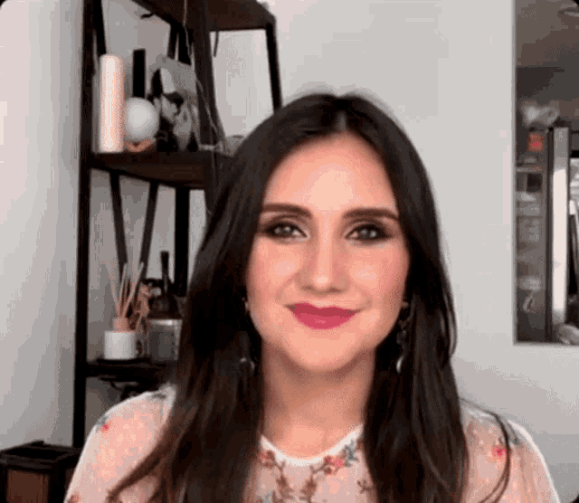 a woman with long dark hair is standing in front of a shelf and smiling at the camera .