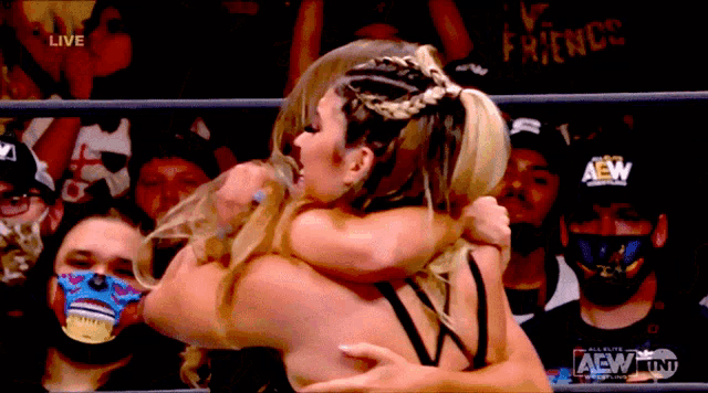 a wrestler is hugging another wrestler in front of a crowd with a aew logo on their hat