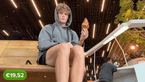 a man is sitting on a bench holding a croissant with a green sticker that says 19,52