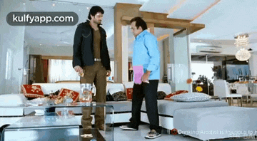 two men are standing next to each other in a living room with a glass table .