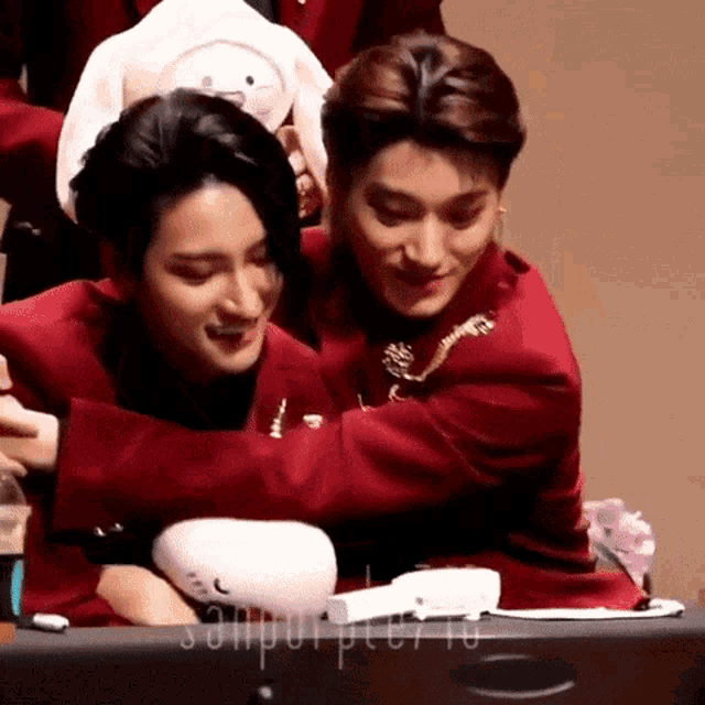 two men in red jackets are hugging each other while sitting at a table .