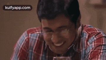 a man wearing glasses and a plaid shirt is laughing while sitting at a table .