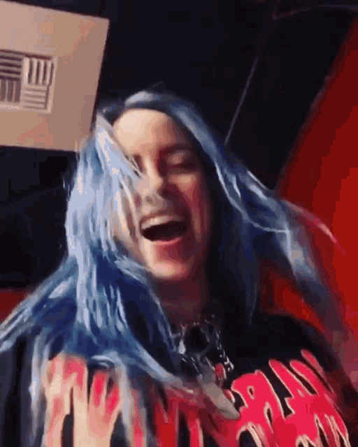 billie eilish has blue hair and is wearing a black shirt with flames on it and laughing .