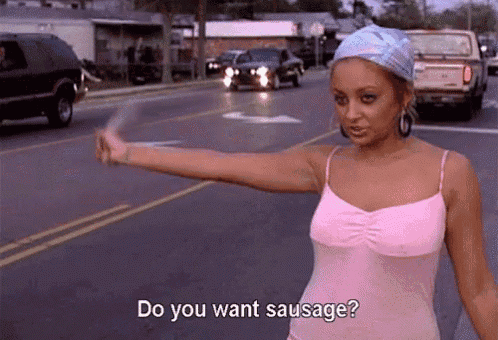 a woman in a pink tank top is standing on the side of a road holding a stick and asking do you want sausage .