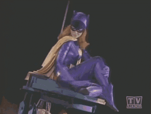 a woman in a purple batman costume is sitting on a table with a tv logo in the background