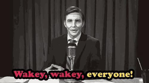 a man in a suit and tie stands in front of a microphone with the words wakey wakey everyone behind him