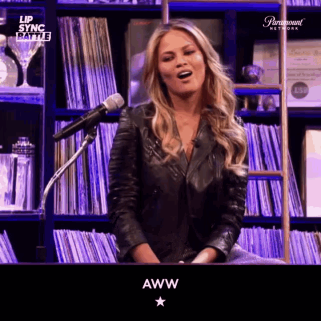 a woman in a leather jacket is singing into a microphone in front of a bookshelf .