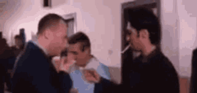 a group of men are standing in a room talking to each other . one of the men is smoking a cigarette .