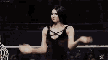a woman in a black tank top is standing in a wrestling ring with her hands folded .