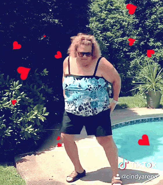 a woman in a blue top and black shorts is standing in front of a pool with hearts around her