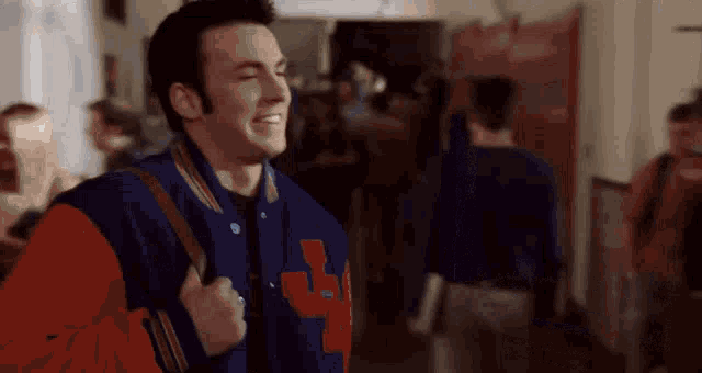 a man wearing a varsity jacket is walking down a hallway with a backpack .
