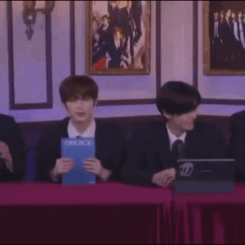 a group of people are sitting at a table looking at a tablet with a picture of a group of people on the wall behind them