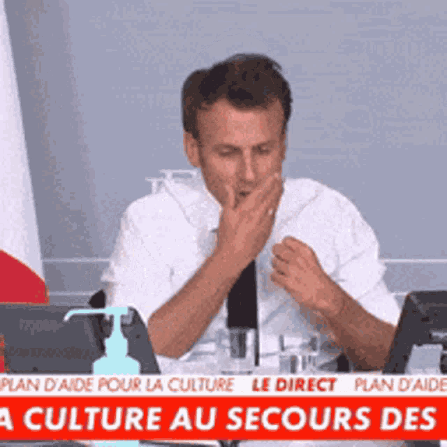a man is covering his mouth while sitting at a desk in front of a sign that says ' culture au secours des '