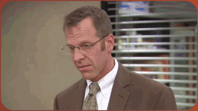 a man wearing glasses and a suit is standing in front of a window with blinds