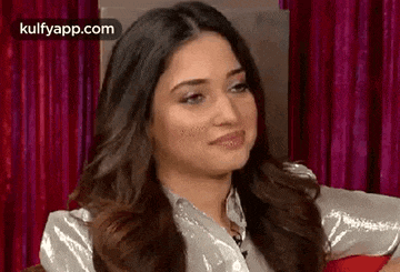 a woman with long hair is sitting in front of a red curtain and making a funny face .