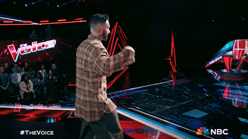 a man in a plaid shirt stands on a stage in front of a nbc logo