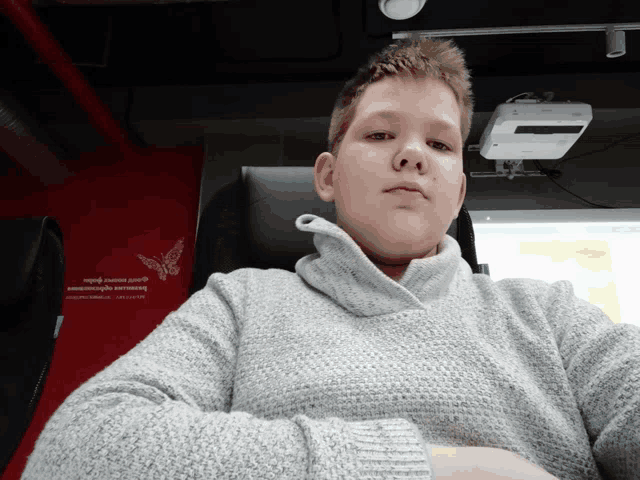 a boy in a grey sweater sits in front of a red wall that says butterfly