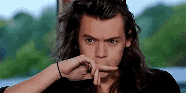 harry styles with long hair is making a funny face with his finger in his mouth .