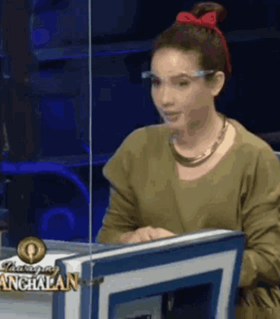 a woman wearing glasses and a red bow in her hair is sitting at a desk in front of a screen .