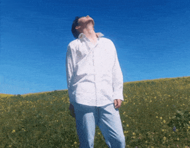 a man in a white shirt stands in a field of flowers looking up at the sky