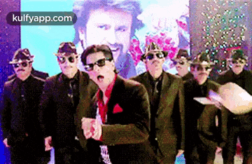 a group of men in suits and sunglasses are dancing in front of a picture of a man in a suit .