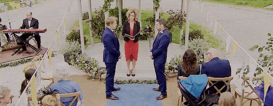 two men are getting married in front of a crowd of people while a man plays a keyboard in the background .