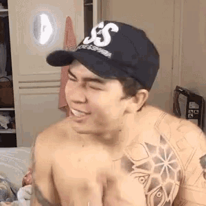 a shirtless man wearing a baseball cap and a tattoo on his arm is laughing .