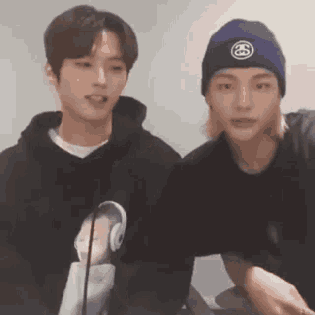 two young men are standing next to each other in a room . one of the men is wearing a beanie and headphones .