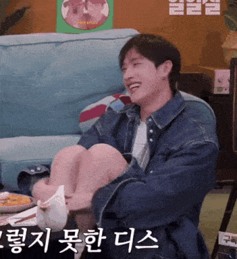 a man in a denim jacket is sitting on a couch with his legs crossed and smiling in korean