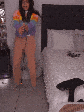 a woman wearing headphones and a rainbow sweater stands in front of a bed