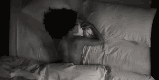 a black and white photo of a woman laying in bed with white sheets .