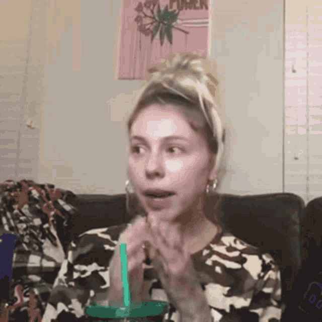 a woman in a camouflage shirt is sitting on a couch drinking from a green cup with a straw .