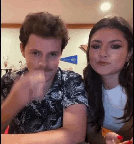 a man and a woman are sitting at a table and the man is pointing at the camera while the woman looks at him .