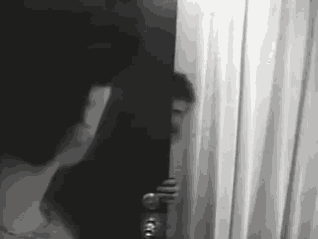 a black and white photo of a man peeking through a curtain